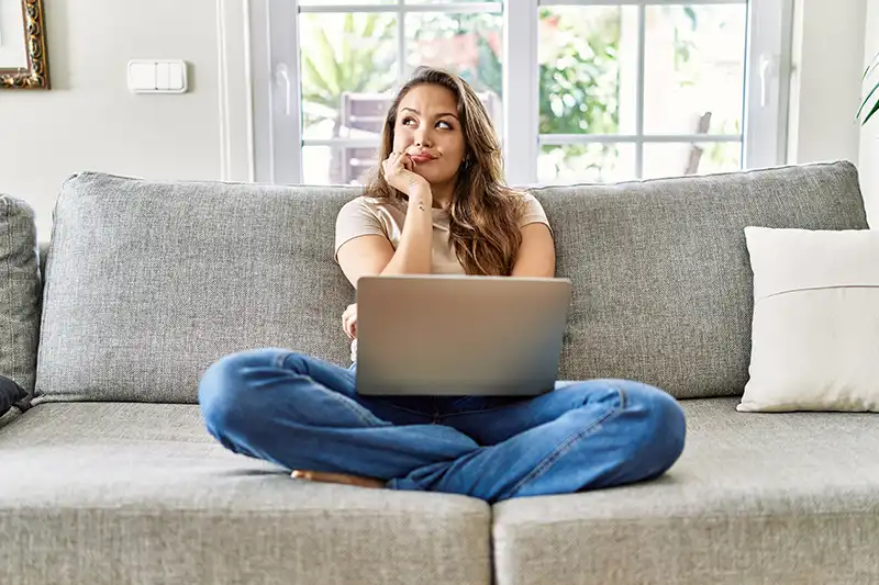 Junge Frau sitzt nachdenklich mit Laptop auf dem Sofa – sie informiert sich über Zahnzusatz-Versicherungen und vergleicht Tarife online.